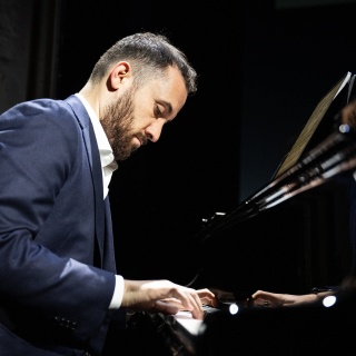 Igor Levit spielt im dunkelblauem Anzug vor schwarzem Hintergrund am Klavierflügel