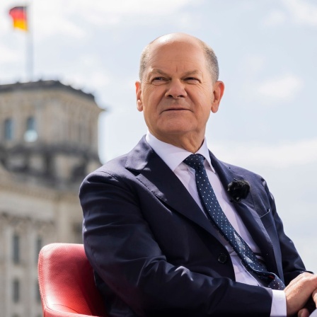 Bundeskanzler Olaf Scholz sitzt vor einem Interview in einem Sessel, im Hintergrund das Reichstagsgebäude.