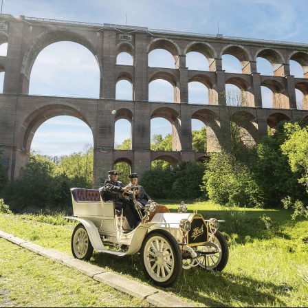 Ein historisches Automobil mit zwei Personen vor einer Bogenbrücke.