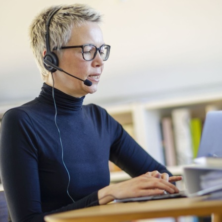 Eine Frau sitzt zu Hause am Schreibtisch. Sie trägt ein Headset und spricht.