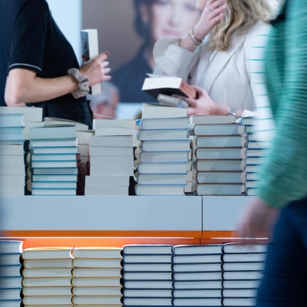 Bücherstapel sind am Stand eines Verlages aufgetürmt. Besucherinnen halten Bücher in der Hand. Der Verlag präsentiert sich in einer Halle der Frankfurter Buchmesse (2024).