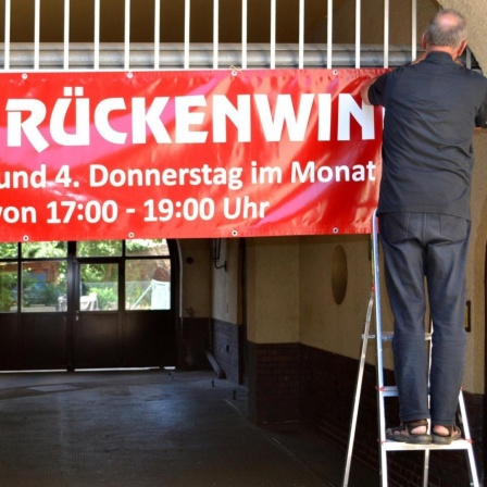 Stefan Friedrichowicz steht auf einer Leiter und hängt ein rotes Banner mit der Aufschrift "Cafe Rückenwind" auf.