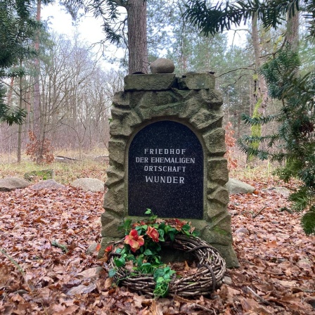 Im Wald steht ein Gedenkstein mit der Aufschrift "Friedhof der ehemaligen Ortschaft Wunder". Davor wurde ein Grabkranz abgelegt.
