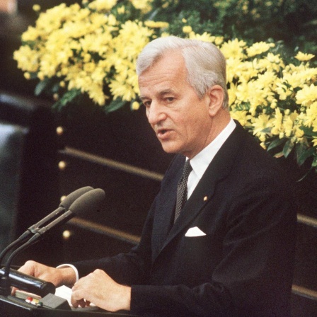 Der deutsche Bundespräsident Richard von Weizsäcker bei seiner vielbeachteten Rede im Bonner Bundestag am 8.5.1985 während der Feierstunde zum Ende des 2. Weltkrieges. Er steht im schwarzen Anzug vor einem gelben Blumenbouquet am Mikrofon.