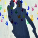 Ein Mann mit einem Kind auf dem Arm und einem an der Hand wirft einen Schatten auf eine mit bunten Handabdrücken bemalte Wand einer Kindertagesstätte.