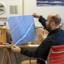 Julius Bockelt sitzt in seinem Atelier am Arbeitstisch und hält eines seiner Bilder in den Händen. Es zeigt hunderte blaue, sehr fein und eng beieinader gezeichnete Linien.