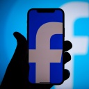 Eine Hand hält ein Smartphone, auf dessen blauem Bildschirm ein kleines weißes "f" zu sehen ist, das Symbol für Facebook.