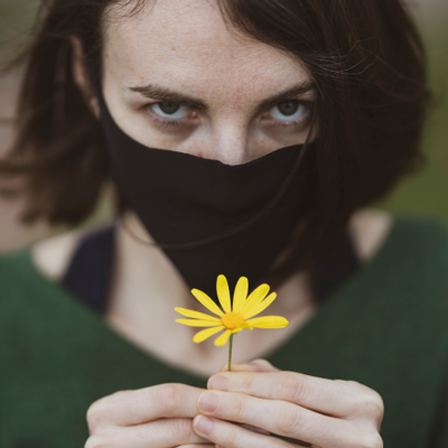 Frau trägt schwarze Maske und hält gelbe Blume in der Hand.