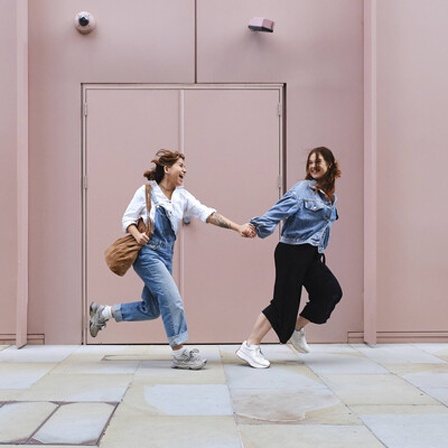 Zwei Frauen laufen Hand in Hand an einem hellrosa Gebäude vorbei