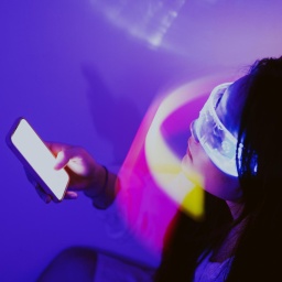 Eine Person trägt eine virtuelle Brille und hält ein Smartphone in der Hand. Die Person sitzt in einem violett ausgeleuchteten Raum.