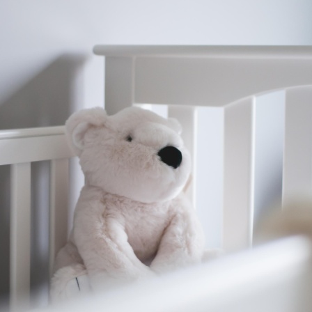 Ein weißer Teddybär sitzt ein einem weißen Kinderbett.