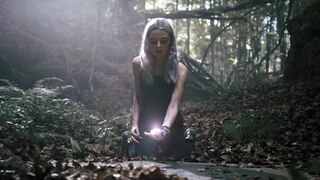 Eine Frau mit silbrigem Haar kniet auf dem Waldboden, in ihrer Hand leuchtet ein Gegenstand