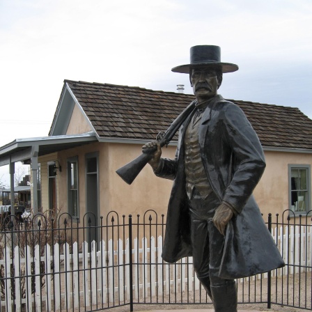 Eine Bronze-Statue des Künstlers Tim Trask erinnert einem Friedhof in Arizona an den Revolverhelden Wyatt Earp. Eine Lebensgroße Figur mit Mantel steht auf einem Sockel und hält ein Gewehr. 
