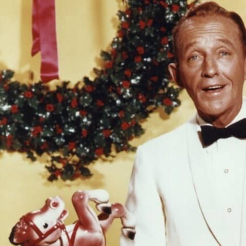 Der Sänger Bing Crosby (&#034;White Christmas&#034;) steht in einem weißen Sakko und Fliege mit einer rot-weißen Weihnachtsmütze neben einem Sack vom Weihnachtsmann vor einem gelben Hintergrund.