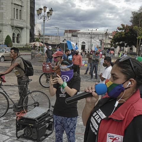 Demonstrierende in Guatemala-Stadt haben sich zum Protest gegen Korruption versammelt, eine Frau in roter Weste spricht in ein Mikrofon, alle tragen Mund-Nase-Masken und halten Abstand voneinander, um sich während der weltweiten Covid-19-Pandemie vor Ans