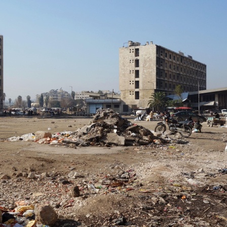 Menschen gehen über einen kargen Platz in Aleppo auf dem sich ein kleiner Schutthügel türmt.