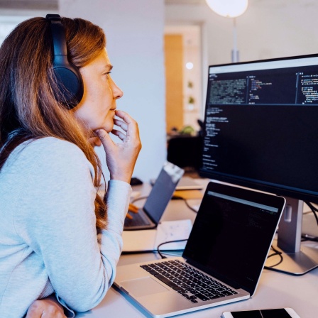Eine Frau mittleren Alters mit schulterlangen Haaren und Kopfhörern sitzt vor einem Computer mit einer Programmier-Oberfläche und grübelt. Sie trägt einen grauen Pullover und stützt das Kinn mit der Hand ab.