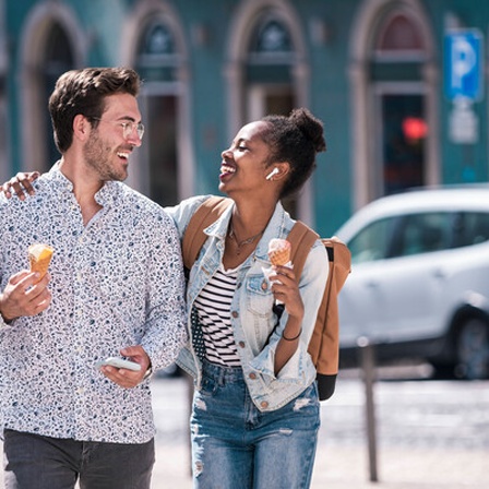 Ein Mann und eine frau essen Eis und schlendern durch eine Stadt. Die Frau legt einen Arm um die Schulter des Mannes.