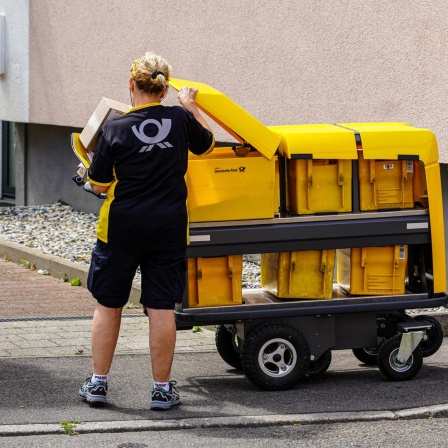 Eine Briefträgerin nimmt Briefe aus ihrem modernen Postwagen der Deutschen Post, um sie in die Briefkästen eines Mehrfamilienhauses einzuwerfen.