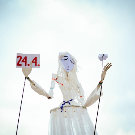 Eine große Marionette mit traurigem Blick, hält Schild mit dem Datum 24.4. in der rechten Hand