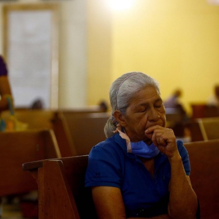 In einer Kirchenbank sitzt allein eine ältere, grauhaarige Frau. Im Hintergrund ist eine jüngere Frau mit langen dunklen Haaren und einem weißen Mundschutz zu sehen.
