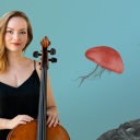 Eine junge Frau in schwarzem Slip-Dress mit offenem blondem Haar schaut direkt in die Kamera. Vor ihr steht ein Cello, auf ihrer Brust ist eine lange, senkrechte Narbe.