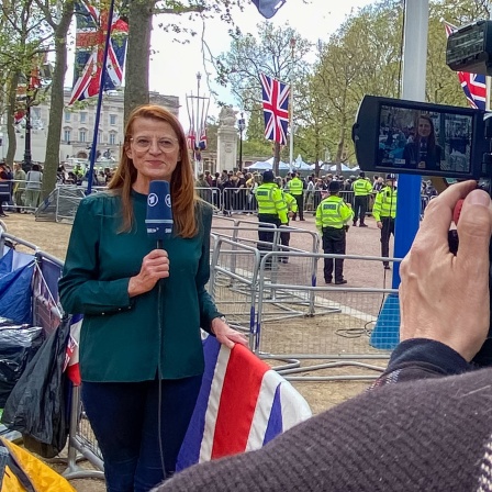 Annelie Malun steht vor britischen Fahnen in London. Sie hält ein ARD-Mikrofon in der Hand. Im Vordergrund rechts ein Mann, der sie filmt.
