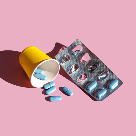 Ein Blister mit blauen Pillen liegt neben einem gelben Becher auf rosa Grund. Vor dem Becher liegen ein paar lose Pillen.