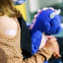 Ein Kind hält einen blauen Dino als Kuscheltier im Arm. An ihrem Arm hat sie ein Impfpflaster.