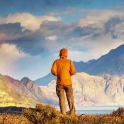 Ein von hinten fotografierter Mensch in orangener Outdoorjacke, der auf ein Bergpanorama blickt.