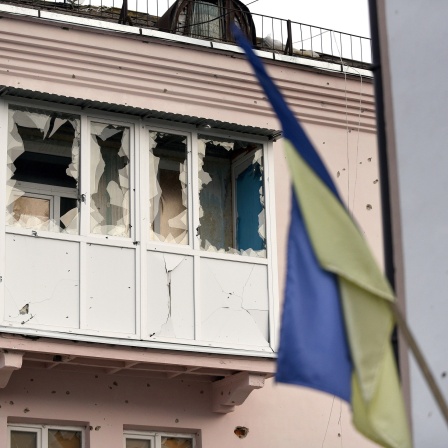 Ein durch russischen Beschuss zerstörter Balkon mit Einschusslöchern und gesplitterten Scheiben in Isjum im Nordosten der Ukraine ist hinter einer ukrainischen Flagge zu erkennen.