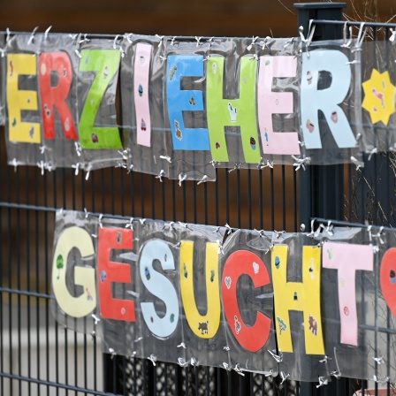 Mit bunten Buchstaben sind die Worte „Erzieher*in gesucht“ geformt, die an einem Zaun einer Kindertagesstätte befestigt sind.