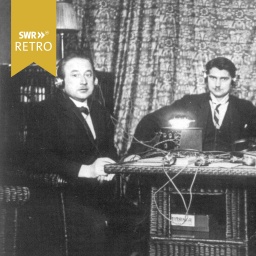 Zwei Männer sitzen an einem Tisch mit Kabeln und hören über Kopfhörer die erste Radiosendung der SÜRAG ab