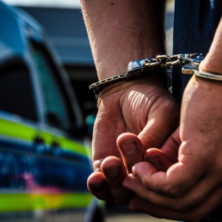 Zwei Hände, die in Handschellen liegen, im Hintergrund zeichnet sich ein Polizeifahrzeug ab.