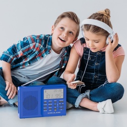 Ein Junge und ein Mädchen sitzen auf dem Boden im Schneidersitz. Vor ihnen steht ein blaues Radio. Das Mädchen hat Kopfhörer auf und der Junge lehnt sich zu ihr herüber und hört mit.
