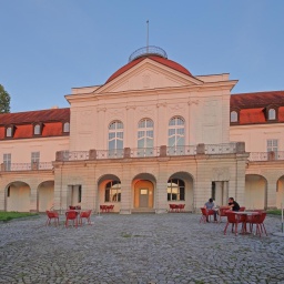 Blick auf das Deutsche Literaturarchiv in Marbach: Das Gebäude wird in rosa von der Sonne angeleuchtet, davor stehen einige Tische und Stühle. 