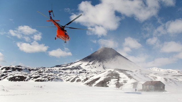 Vulkan mit schwarzer Spitze in weißer Schneelandschaft unter blauem Himmel mit wenigen weißen Wolken, vorne ein roter Hubschrauber, der auf den Vulkan zufliegt, und eine kleine blassblaue Hütte