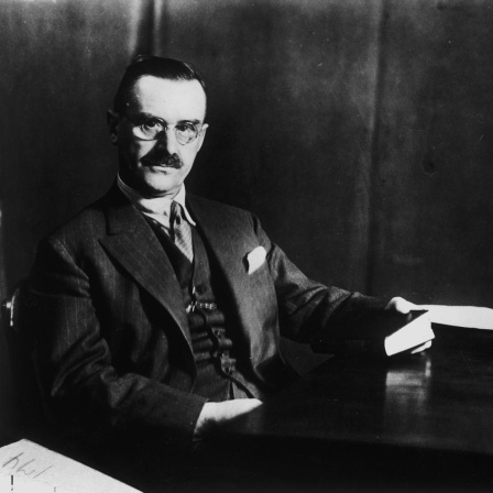 Der Schriftsteller Thomas Mann sitzt an einem Schreibtisch, in seiner Hand ein Buch. Rechts auf dem Schreibtisch steht ein Mikrofon.