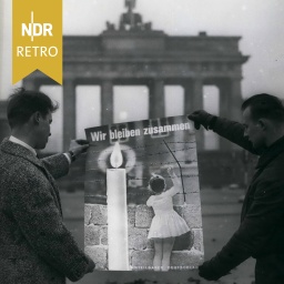 Zwei Männer halten ein Plakat des „Kuratorium Unteilbares Deutschland“ vor das Brandenburger Tor in Berlin. Auf dem Plakat ist der Schriftzug „Wir bleiben zusammen“ abgebildet. November 1961