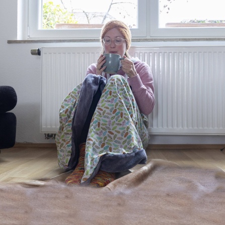 Frau sitzt, in eine Decke gewickelt, vor einem Heizkörper und bläst in eine Tasse.