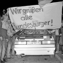 Menschen stehen an der innerdeutschen Grenze freuen sich. Über einem Auto wird ein Banner mit der Aufschrift „Wir grüßen alle Bundesbürger“ gehalten.