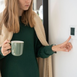 Frau mit Decke um die Schultern und Tasse in der Hand stellt das Thermostat ein: Menschen nehmen Kälte unterschiedlich wahr. Das hat mit dem Körperbau zu tun. Aber das Kälteempfinden hängt auch von äußeren Umständen ab. Die kann man beeinflussen.