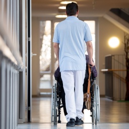 Ein Pfleger schiebt eine Bewohnerin eines Pflegeheims in einem Rollstuhl über den Flur.