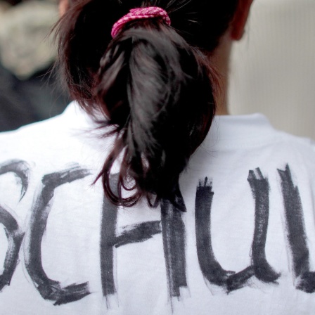 Ein Mensch mit Zopf trägt ein weißes T-Shirt auf dessen Rücken das Wort "Schuld" geschrieben steht.