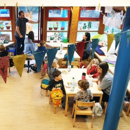 Ein Raum in einer Kindertagesstätte mit vielen Kindern und drei Erziehern, die an vier Tischen sitzen. 