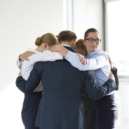 Fünf Männer und Frauen in Business-Kleidung bilden umarmend einen Kreis, eine Frau schaut weg