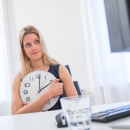 Frau hält am Schreibtisch große Uhr: Mütter und Väter haben ein Recht, nach der Elternzeit tageweise oder halbtags zu arbeiten - so manches Mal landen sie in Teilzeit aber nur auf unattraktiven Positionen.