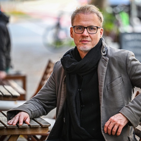 Falk Richter sitzt an einem Tisch in einem Biergarten. Seine Hand liegt auf dem Tisch, er trägt Mantel und Schal sowie eine schwarz umrandete Brille.