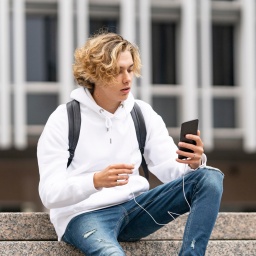 Jugendlicher situt mit Smartphone auf einer Treppe vor einem Schulgebäude