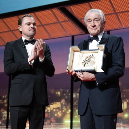 Leonardo Di Caprio und Rober Di Niro auf einer Bühne in Cannes. Di Caprio applaudiert mit einem stolzen und leicht gerührten Gesichtsausdruck. Di Niro hält eine geöffnete Schatulle in den Händen, in der die „Goldene Palme“ (Palme d'Or) zu sehen ist – die höchste Auszeichnung der Internationalen Filmfestspiele von Cannes. Der Hintergrund zeigt eine große Fensterfront mit Blick auf eine stilisierte Stadtansicht bei Sonnenuntergang oder in der Dämmerung. Die Szene ist stark beleuchtet und vermittelt eine festliche, respektvolle Atmosphäre. 